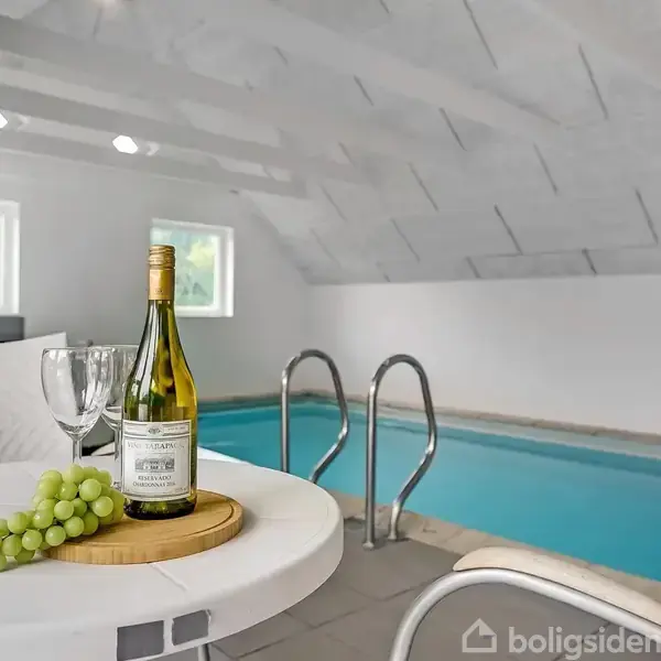 En lille indendørs swimmingpool er omgivet af hvide vægge med tagvinduer. Foran ses et rundt bord med en vinflaske, to glas og grønne druer på en skærebræt.