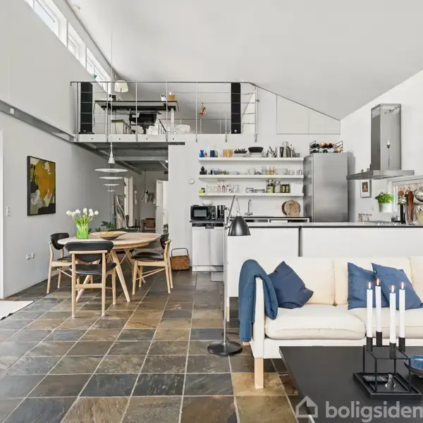 Lyst, moderne stue med åbent køkken og spiseområde. Sofa står foran et sofabord med lysestager. Over spisebordet hænger en lampe. Ovenpå er der en hems.