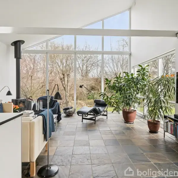 Stue med skråloftet og store vinduer. En loungestol står tæt ved planter i potter, mens en pejs er i brug. Rummet har flisegulv, sofa, og udsigt til træer.
