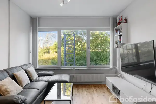 En sort lædersofa står i en stue foran store vinduer, der viser en grøn have udenfor. En sort tv-skærm hænger på væggen over en lav, hvid enhed.