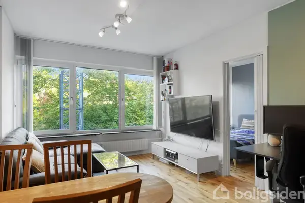 Stue med spisebord i forgrunden og sofa langs vinduet, der viser grønne træer udenfor. Fladskærm på væggen, reol ved siden af og en åbning til et andet værelse.