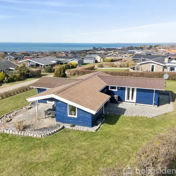 Et blåt hus med brunt tag står på en velplejet græsplæne. Huset har en overdækket terrasse og er omgivet af flere lignende huse. Havet kan ses i baggrunden.
