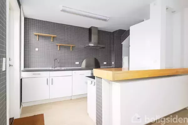 Køkken med hvide skabe og grå fliser på væggen. Et stålvaskarmatur ses ved arbejdsbordet og en emhætte er placeret over kogepladen. To træhylder hænger på væggen.