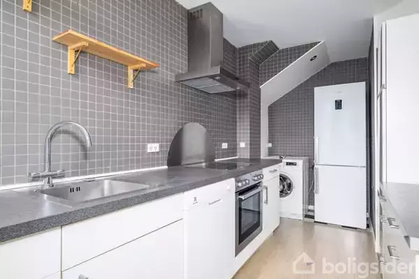 Hvidt køkken med grå flisevæg. Der er en vask, komfur og emhætte, samt en vaskemaskine og køleskab i baggrunden. En træhylde er monteret på væggen.