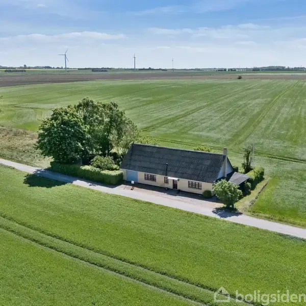 Lille hus ligger stille ved en landevej omgivet af åbne marker og træer. I baggrunden ses flere vindmøller, og himlen er klar med spredte skyer.