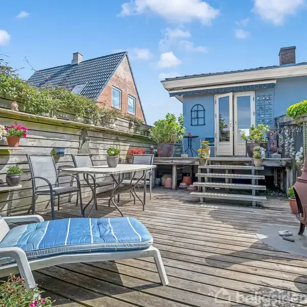Solstol står på en træterrasse. Bord med stole og krukker omkranser terrassen. En trappe leder til et hus med en glasdør. Blomster og en pejs skaber en hyggelig atmosfære.