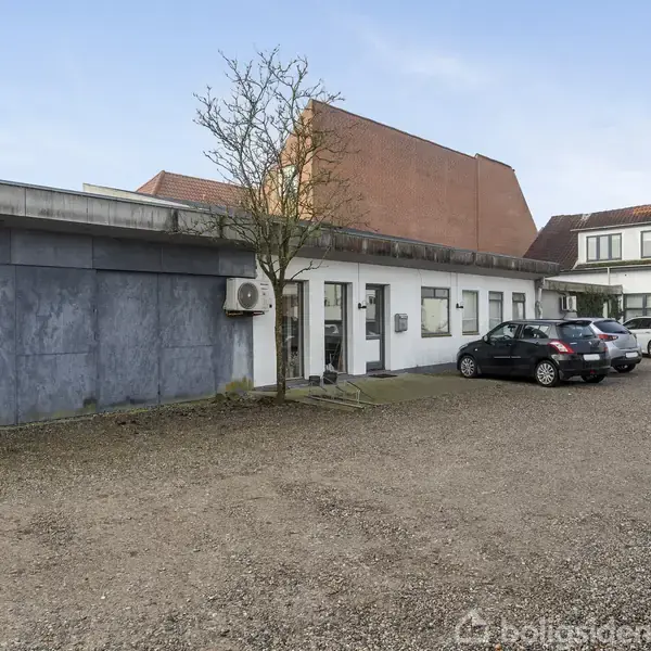 En parkeringsplads ved siden af en lav bygning med et skilt "00" på væggen, hvor biler holder parkeret foran bygningen i et grusbelagt område med spredte træer.
