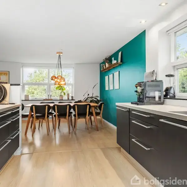 Køkken-alrum med sorte skabe og opvaskemaskine på højre side; stort spisebord med seks stole i baggrunden; grøn væg med hylde og kunstværker; store vinduer; grøn udsigt.