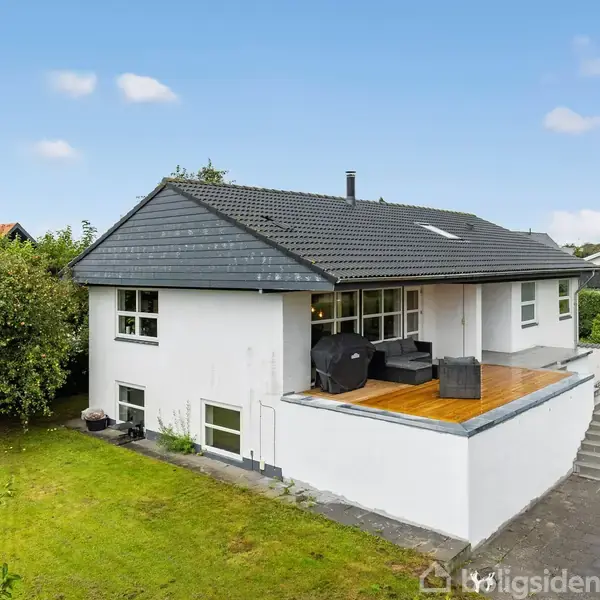 Hvidt hus med mørkt tag står i grøn have. Der er en træterrasse med møbler. Omgivet af buske og træer, med en asfalteret indkørsel til højre.