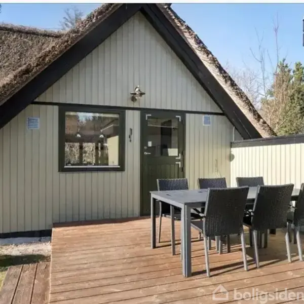 Et stråtækt hus med gavl i træ er hovedobjektet. En have indeholder et spisebord med seks stole på en terrasse af træ. Omgivet af træer og buske.