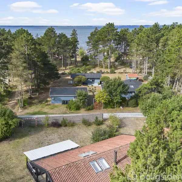 Et hus med rødt tegltag står blandt grønne træer tæt ved en kyst. Flere små huse ses i baggrunden, omgivet af skov, med havudsigt i horisonten.