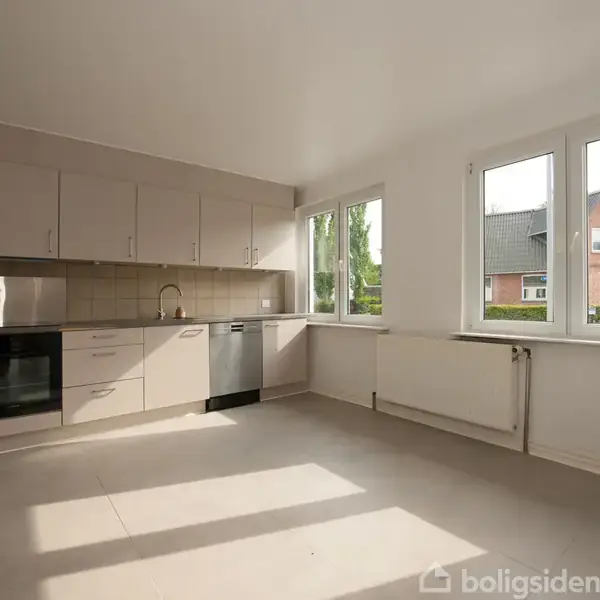 Køkken med lysegrå skabe og indbygget ovn, opvaskemaskine og vask. Sollys strømmer ind gennem store vinduer. Udenfor ses en have og et rødt hus.