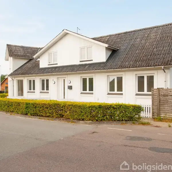 En hvid, etplans villa med mørkt tag står stille i et villakvarter. Omkranset af grønne hække og en træhegn ved vejen, med andre huse i baggrunden.