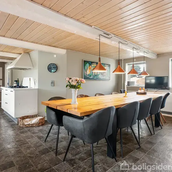 Langt træbord med otte stole omgives af en moderne, lys spisestue. Bordet har en vase med blomster. Bagved ses et åbent køkken med en stor køleskab og ovn. Pendellamper hænger fra loftet.