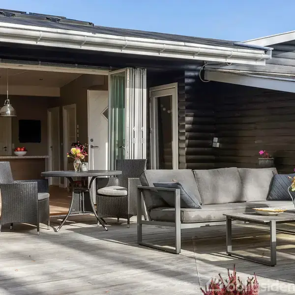 Terrassemøbler står på en træterrasse foran et træhus. En sofa og et bord er placeret udendørs, omgivet af stole og blomster. Indenfor ses et åbent køkken med hængende lamper.