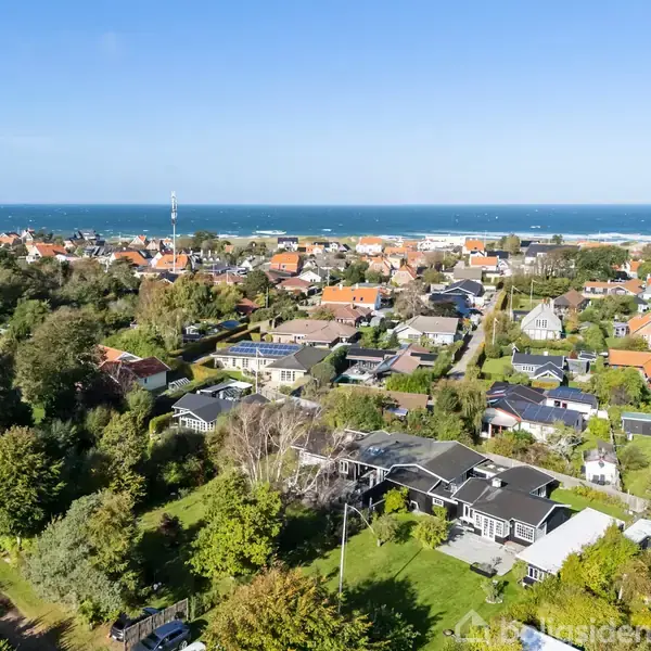 Luftfoto af et kystnært boligområde med huse omgivet af træer og haver. I baggrunden ses havet og en klar himmel.