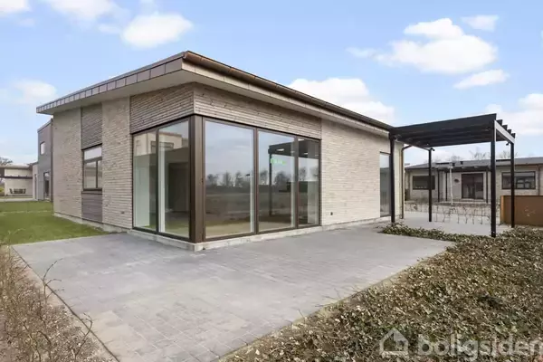 Et moderne hus med store vinduespartier står på en flisebelagt terrasse. Omkring er der en grøn plæne og himlen er let overskyet.