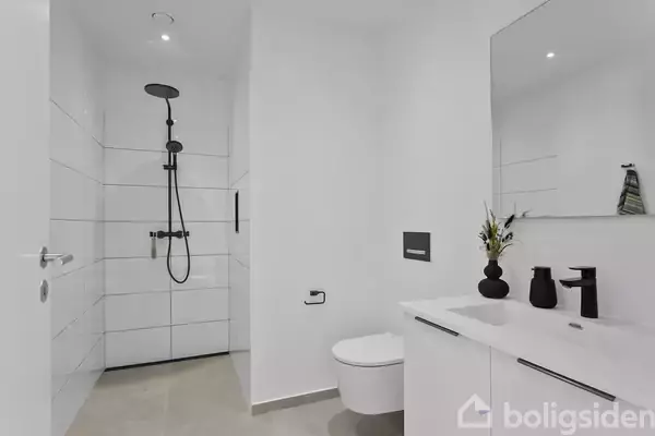 Et moderne badeværelse med hvidt interiør, der indeholder en bruseniche med sort brusearmatur, et væghængt toilet og en dobbelt håndvask med spejl. En sort vase står på vasken.