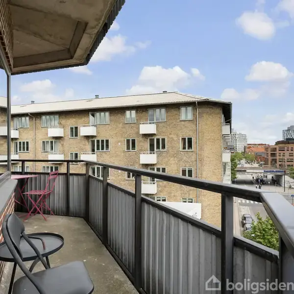 Balkon med to stole og bord, oppe i en beboelsesejendom, udsigt over bymiljø med flere bygninger.