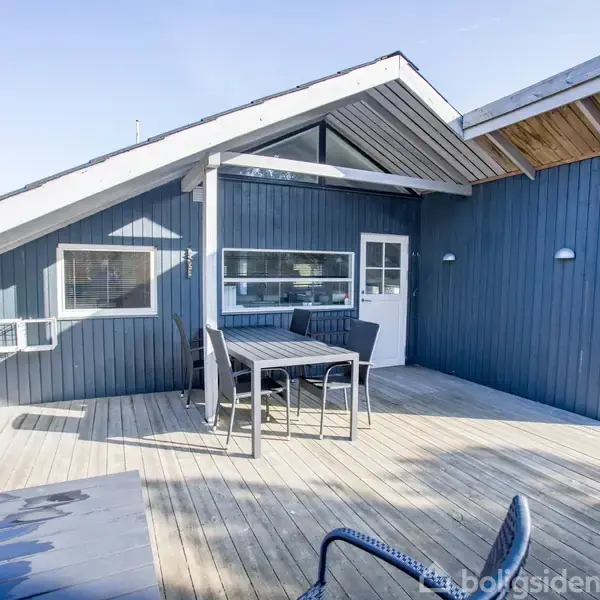Et moderne sommerhus i træ med blå facade. En stor terrasse med bord og stole står ud for huset, omgivet af træer og klar himmel.