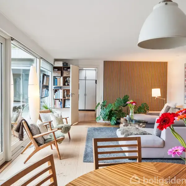 Stue med sofa og lænestol ved siden af et stort vinduesparti, der fører til en terrasse. Rundt spisebord med blomster i forgrunden, væghylde og planterr ved siden af.