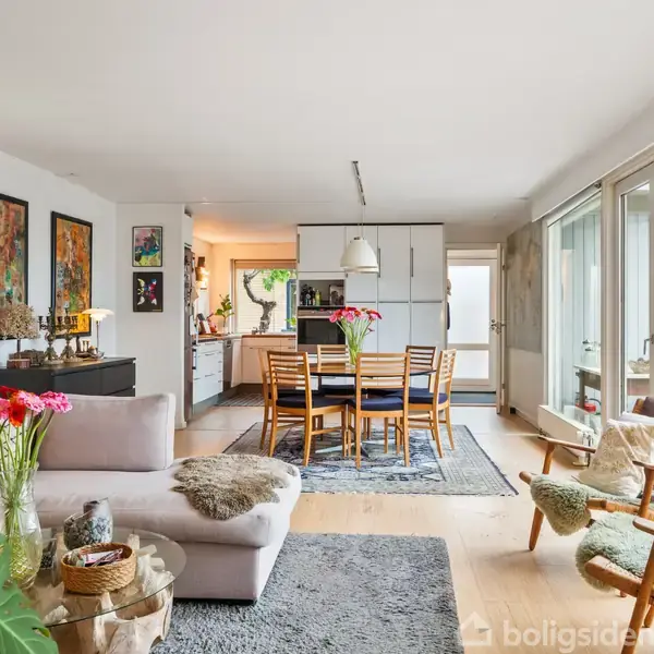 Stue med lys sofa og blomster på sofabordet; spisebord i baggrunden med stole omkring; store vinduer til højre slipper lys ind, hvilket skaber en lys og indbydende atmosfære.