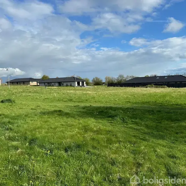 Grønt græsfelt breder sig foran to moderne, lave bygninger under en delvist skyet himmel. Bygningernes enkle konstruktion omkranses af træer og buske i en landlig setting.