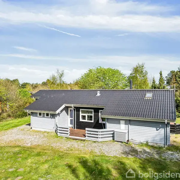 Gråt, enkelt hus med sort tag står stille omgivet af en grøn have og træer. Grus indgangsvej fører til hoveddøren, beliggende under en blå himmel med lette skyer.