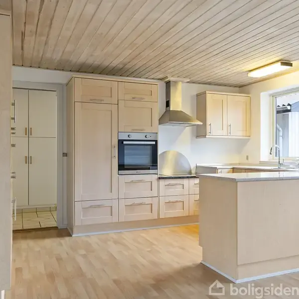 Et moderne køkken med lyse træskabe, en indbygget ovn, og en stor emhætte. Der er et køkkenbord ved et vindue, som giver naturligt lys. Golvet er af lyst træ.
