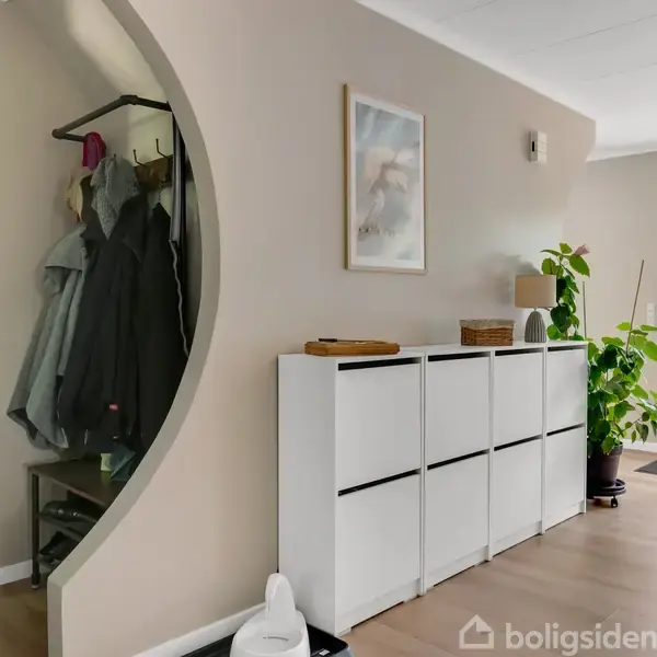 En entré med en hvid skoskab op ad væggen, over hvilken der hænger et billede. En åben garderobe med jakker ses til venstre. En plante og dør ses til højre.