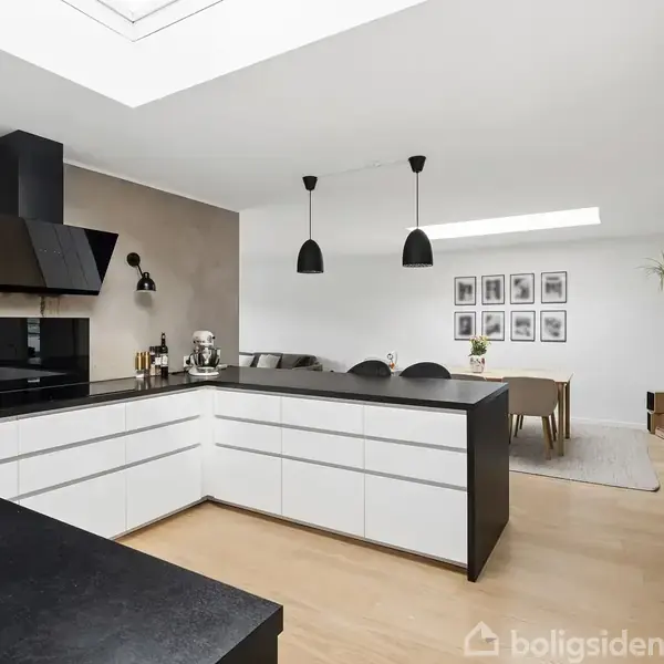 Et moderne køkken med en L-formet sort og hvid bordplade, sorte lamper hængende over. I baggrunden ses en lysegrå væg med indrammede billeder og et spisebord med stole.