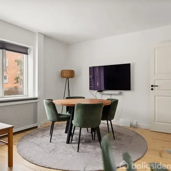 Et rundt spisebord med fire grønne stole står på et gråt tæppe. Et tv er monteret på væggen. Et skilt i vinduet læser "Til Salg" med "danbolig."