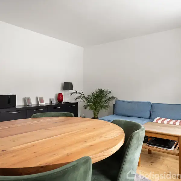 Rundt spisebord med grønne stole står i en stue. Blå sofa med pudestripe, træbord, sorte møbler og planter i nærheden. Væglampe hænger i hjørnet. Ingen tekst fundet.