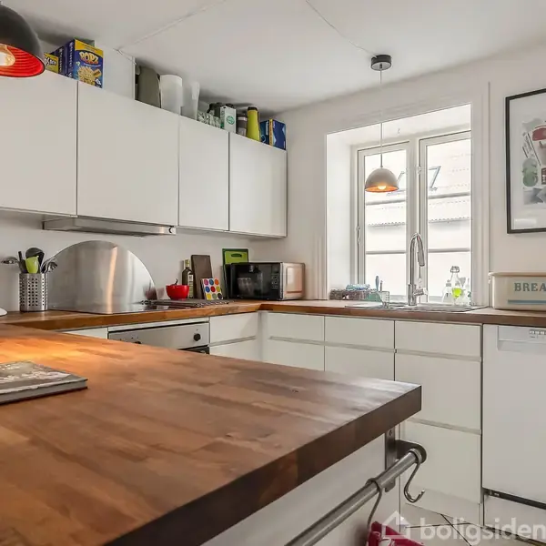 Et moderne køkken med hvide skabe og træbordplader. En kande og brødkasse ses på bordet, mens diverse køkkenredskaber og små apparater står på skabene. En stor vindue lader lys komme ind.