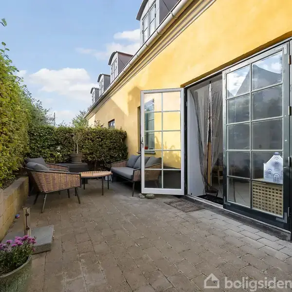 Terrasse med møbleringer, inklusive kurvestole og bord, ligger op ad en gul murstensbygning. Åben glasdør fører ind i huset. Omringet af grønne buske og blomster i krukker.