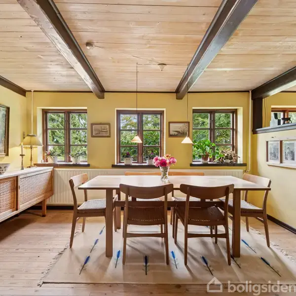 Et spisebord med seks stole står i et lyst rum med gule vægge og træloft. Store vinduer lader naturligt lys ind, og et skab står til venstre med billeder på væggen.
