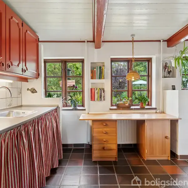 Et køkken med røde skabe, en vask, og stribede gardiner under bordpladen. Tre vinduer med udsigt til have. Der hænger en lampe over et lille træskrivebord.
