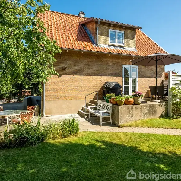 En rødstenet hus med skråtag og en lille terrasse med parasol. Terrassebord og stole står nær grønne planter i en have med græsplæne i solskin.