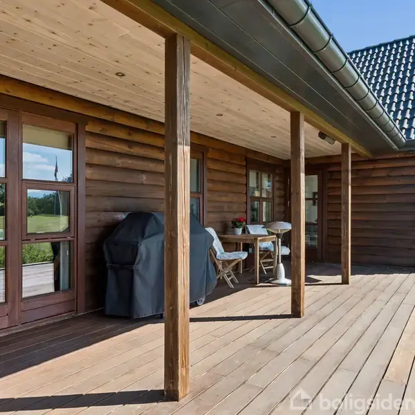 En træterrasse langs et bjælkehus har et bord med to stole og en grill. Omgivet af grønne buske og marker under en klar himmel.
