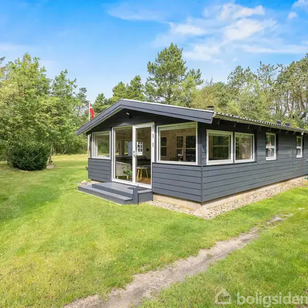 Et gråt sommerhus står stille i en grøn have omgivet af træer. Stien foran huset fører til en indgang med glaspartier.