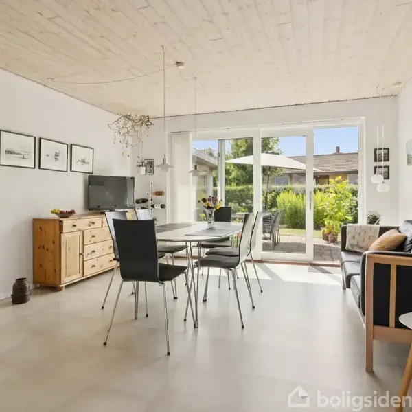 Spisestue med sort spisebord og stole, en trækommode med tv, og en glasdør åbner ud til en have. Lyse vægge og loft, dekoreret med billeder, gulvet er lyst.