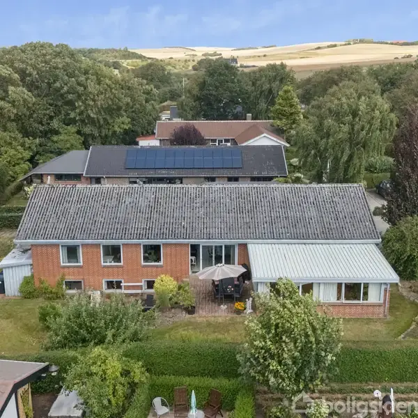 Et rødt murstenshus med grått tag ses foroven. Solpaneler er på taget. Huset er omgivet af en grøn have med træer og buske. Der er landskab og andre huse i baggrunden.
