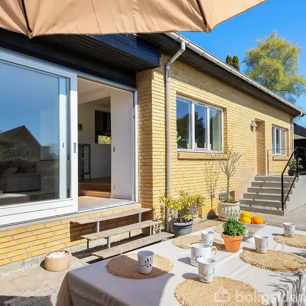 Et gult murstenshus har en terrasse med bord dækket af kopper og tallerkener. Et stort parasol skaber skygge, mens trapper fører op til en dør. Spredte planter omkranser.