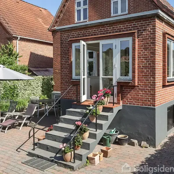 Murstenshus med trappetrin dekoreret med blomsterkrukker fører til en glasdør. Omkringliggende terrasse indeholder liggestole og parasol, flankeret af grøn hæk og andre rødstenshuse.
