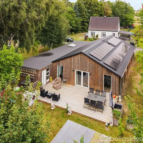 Et moderne træhus med solpaneler på taget står på en grøn grund. Foran huset er en træterrasse med havemøbler. I baggrunden ses et andet hus omgivet af træer.