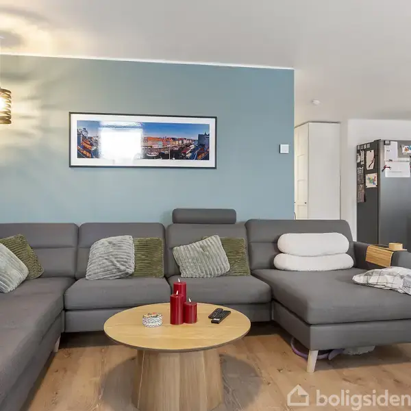 Grå hjørnesofa står i en stue med en lyseblå væg og kunst på væggen. Rundt sofabord med røde og sorte genstande foran sofaen. Køkken med stål-køleskab i baggrunden.