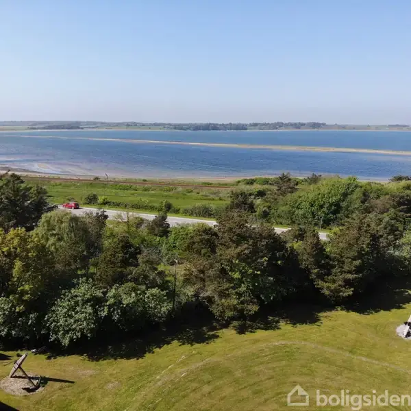 En grøn have med spredte træer strækker sig mod en vej, bagved hvilken en bred flod eller fjord breder sig ud med fjerne, lavtliggende landskaber i horisonten.