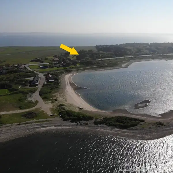 Gul pil peger mod et hus ved kysten, omkranset af vand og strand i et naturskønt område med marker og spredte bygninger.