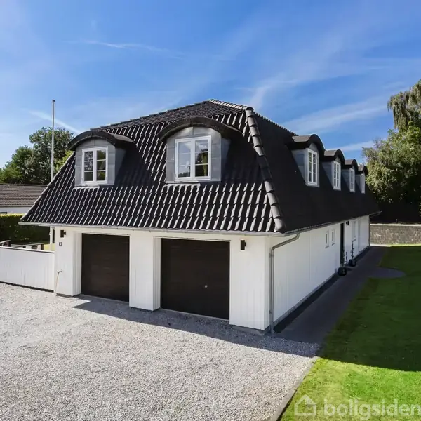 To-garagebygning med sort, mansardtag og hvid facade står på en grusbelagt indkørsel. Omkring er der grønne plæner og træer, under en klar blå himmel.