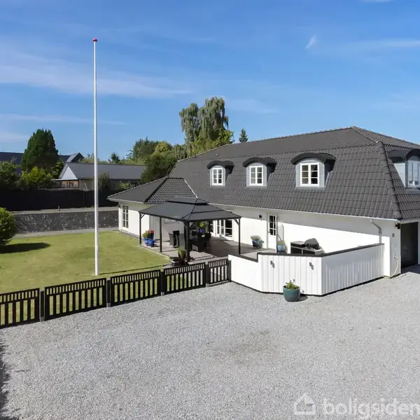 Et hvidt hus med sort tegltag står ved en grusbelagt indkørsel. På verandaen er der havemøbler. En flagstang og en velplejet græsplæne omkranser huset.